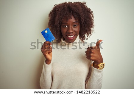 Young african afro woman holding credit card standing over isolated white background happy with big smile doing ok sign, thumb up with fingers, excellent sign