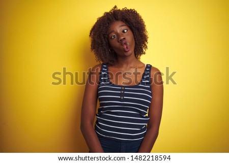 Young african afro woman wearing striped t-shirt over isolated yellow background making fish face with lips, crazy and comical gesture. Funny expression.