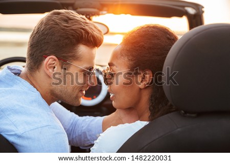 Image of lovely multiethnic couple man and woman kissing while siting in convertible stylish car by seaside at sunset
