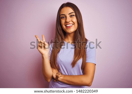 Young beautiful woman wearing casual t-shirt standing over isolated pink background smiling with happy face winking at the camera doing victory sign. Number two.