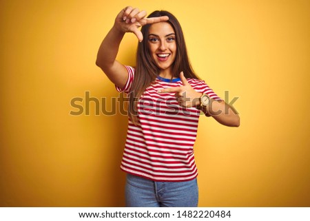 Young beautiful woman wearing striped t-shirt standing over isolated yellow background smiling making frame with hands and fingers with happy face. Creativity and photography concept.