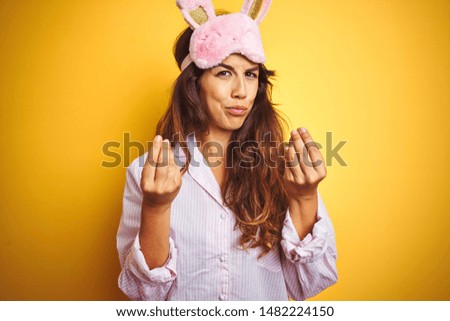Young woman wearing pajama and sleep mask standing over yellow isolated background doing money gesture with hands, asking for salary payment, millionaire business