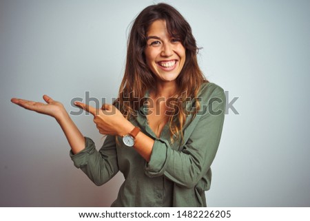 Young beautiful woman wearing green shirt standing over grey isolated background amazed and smiling to the camera while presenting with hand and pointing with finger.