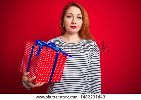 Young beautiful redhead woman holding birthday gift over red isolated background with a confident expression on smart face thinking serious