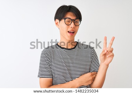 Chinese man wearing glasses and navy striped t-shirt standing over isolated white background smiling with happy face winking at the camera doing victory sign. Number two.