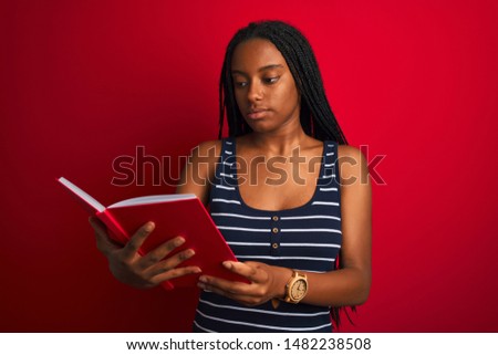 Young african american student woman reading book standing over isolated red background with a confident expression on smart face thinking serious