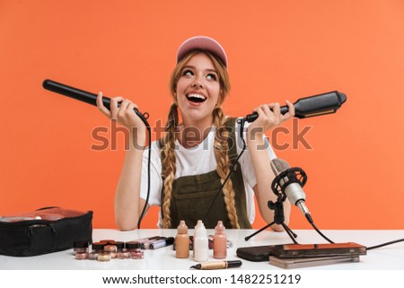 Image of young girl holding hair straighteners while recording blog broadcast about new cosmetic products isolated over orange wall