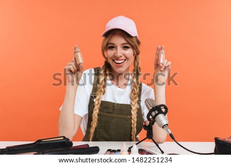 Image of adorable girl recording blog broadcast with microphone about new cosmetic products isolated over orange wall in studio