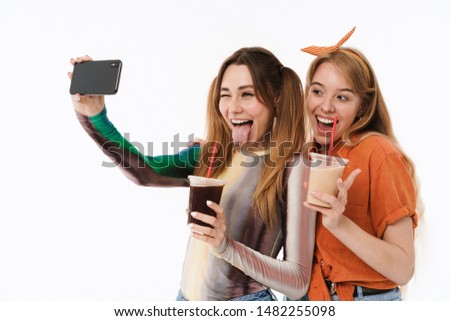 Full length portrait of two caucasian girls wearing casual clothes taking selfie photo and holding plastic cups isolated over white background