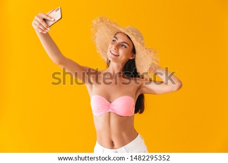 Attractive cheerful young girl wearing bikini standing isolated over yellow background, taking a selfie