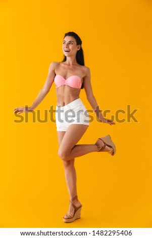 Full length of an attractive cheerful young girl wearing bikini standing isolated over yellow background, posing