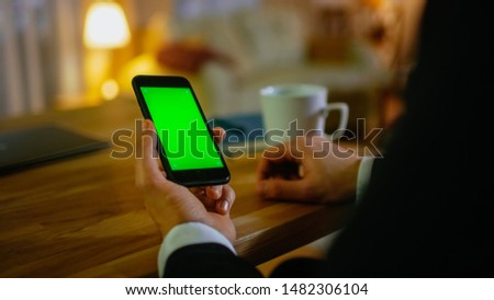 At Home Man Sits at His Desk and Uses Smartphone with Green Screen on It. He Drinks from a Cup. His Apartment is Done in Yellow colours and is Warm.