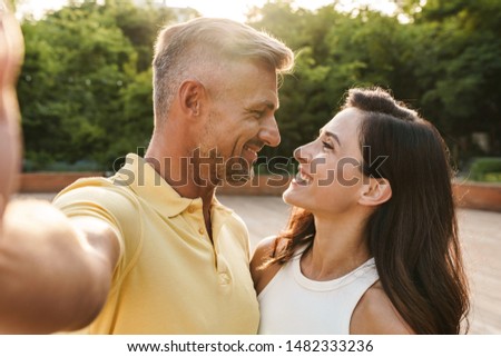 Portrait of joyful middle-aged couple man and woman taking selfie photo and hugging while walking in summer park