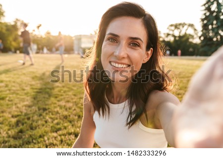 Image of smiling middle-aged woman taking selfie photo and looking at camera while sitting on grass in summer park