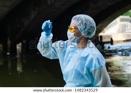 female ecologist or epidemiologist takes test-tube water analysis in a city river under an old bridge Royalty-Free Stock Photo #1482343529
