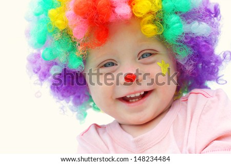 A cute, smiling baby boy is dressed up in a clown costume with colorful wig and clown make up face paint