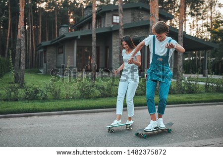 Happy young couple having fun outdoors. Teenage girl and boy are holding hands while skateboarding on the background of beautiful houses.
