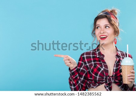 Cute charming young woman in retro clothes is drinking a cocktail from a straw posing against a blue background with copy space and looking to the right. Concept of links and information.