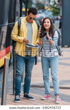 Excited woman with digital camera near bi-racial friend with map and backpack