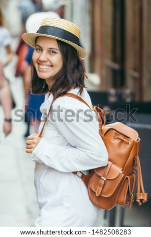 woman in white clothes in straw hat and brown leather backpack walking by city street of venice