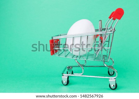 A small copy of a supermarket basket on a green background. Inside is a white egg. The concept of shopping, grocery stores, discounts, promotions.