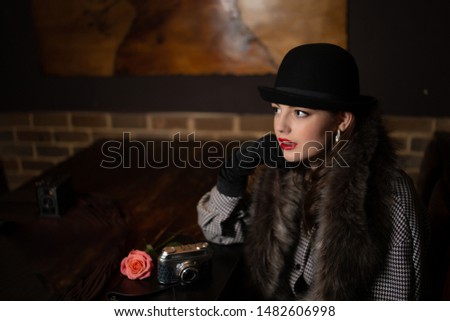 Portrait of a beautiful young girl at an old wooden table in a bar, in retro clothes, a bowler hat with a retro camera and a rose, looking thoughtfully to the side