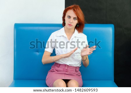 Photo Portrait of a cute woman with bright red hair in a white T-shirt and pink skirt on a black and white background in the studio. Sits on a blue sofa, Talks in front of the camera with emotions.