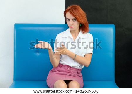 Photo Portrait of a cute woman with bright red hair in a white T-shirt and pink skirt on a black and white background in the studio. Sits on a blue sofa, Talks in front of the camera with emotions.