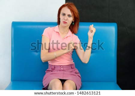 Photo Portrait of a cute woman girl with bright red hair in a peach t-shirt and pink skirt on a black and white background in the studio. Sits on a blue sofa, Talks in front of camera with emotions.