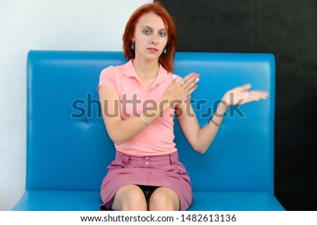 Photo Portrait of a cute woman girl with bright red hair in a peach t-shirt and pink skirt on a black and white background in the studio. Sits on a blue sofa, Talks in front of camera with emotions.