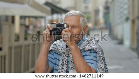 Senior male tourist exploring town and makes a photo with retro photo camera while traveling in Lviv, Ukraine. T-shirt and sweater in his neck. Vacation concept