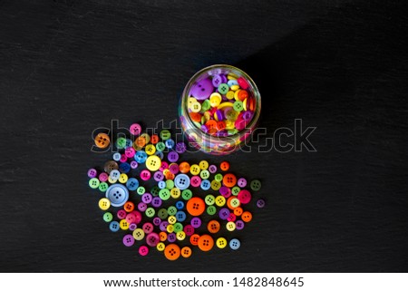Top view of jar of buttons spilled out onto a black background; colorful buttons poured out of clear glass jar