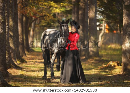 woman with retro costume (Victorian dresses) and retro hat stands next to a black horse in the autumn forest