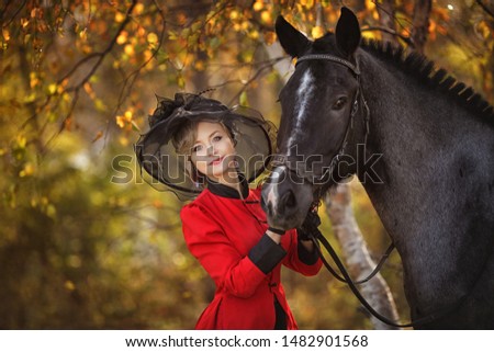 woman with retro costume (Victorian dresses) and retro hat stands next to a black horse in the autumn forest