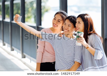 Young Asian people taking selfie photos together inside the glass building. Three beautiful women having fun taking pictures outdoors in blurred background