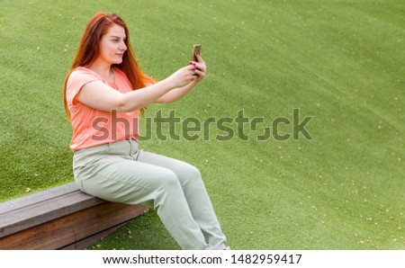 Outdoor portrait of beautiful ginger girl taking a selfie. Happy young woman using smart phone outdoors, girl sitting on a bench in the park.