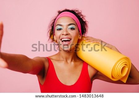 Beautiful african girl with fitness mat posing with smile. Studio shot of cute black lady taking selfie on pink background.