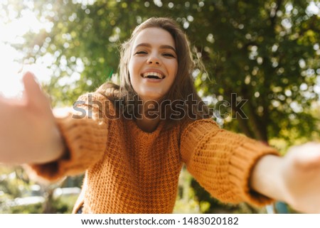 Pleasant young woman taking selfie on blur nature background. Blithesome girl in knitted sweater having fun in park.
