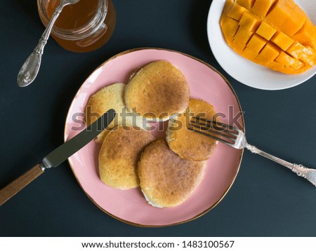 Pancake breakfast on a pink plate with knife and fork. jar of honey with spoon, mango on a white saucer