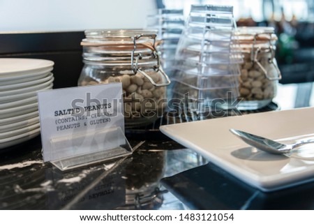 Jars of salted peanuts with a sign warning abouit celery, peanuts and gluten allergens ingredients at a airport business lounge hotel bar.