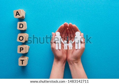 cropped view of woman holding paper cut gay family near blocks with adopt lettering on blue background