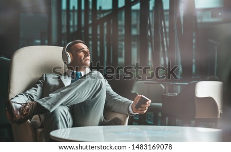 Portrait of relaxed middle-aged businessman in suit having a reast, sitting back in armchair and looking at the window with a remote control in his hand. Horizontal shot