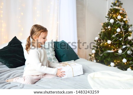 Cute little girl sitting on a child's bed in the background of Christmas tree in bright interior of the house 