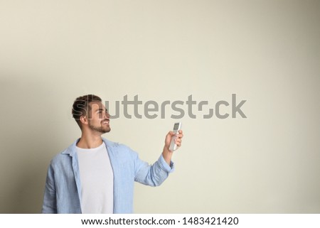 Happy young man operating air conditioner with remote control on beige background. Space for text
