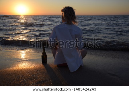Young girl on the sea beach with a bottle of champagne and a glass, the blonde is drinking champagne at the sea. woman is sitting on the sand at the beach.