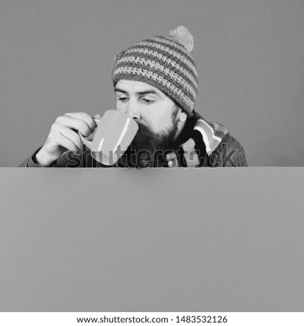 Autumn and hot drink season concept. Hipster with beard and busy face drinks warm tea or coffee. October and November beverage idea. Man in warm hat holds brown cup on green background, copy space