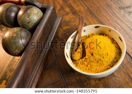 Ceramic bowl filled with yellow curry powder.  There is a wood spoon in the bowl and a decorative wooden tray pictured as well. The bowl is shot on a wood surface.