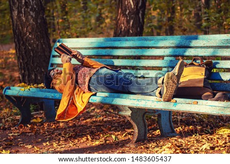 Young stylish hipster girl lying on the bench in the autumn park and reading electronic book