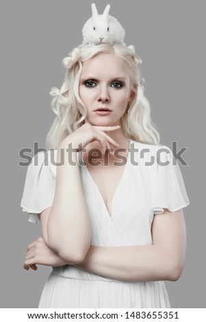 Fashion portrait of albino blond girl with white skin and blue eyes in elegant dress posing with cute little rabbit on gray background in studio