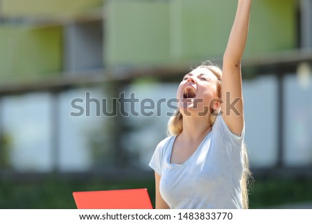 Excited student with laptop celebrating success raising arm outdoors in an university campus
